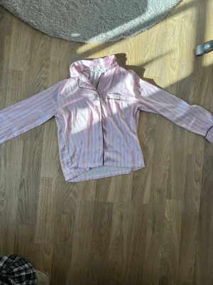 Randig rosa pyjamas med svarta detaljer - Säljer en tvådelad pyjamas med långärmad skjorta och byxor i ljusrosa och vita ränder. 