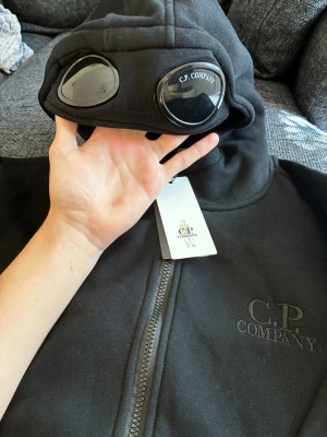 Svart hoodie med goggles från C.P. Company - Säljer en svart hoodie från C.P. Company med deras ikoniska goggles i huvan. Hoodien har dragkedja framtill och broderad logga på bröstet. Tillverkad i mjukt material som känns skönt mot huden. Perfekt för dig som vill sticka ut med en unik streetwear-look. Denna hoodie är i storlek XL men skulle personligen vilja söga att den sitter som en vanlig L