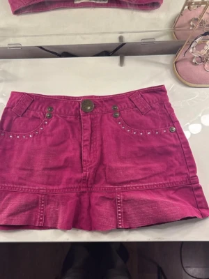 Rosa kortkjol från Guess Jeans - Säljer en snygg rosa kortkjol från Guess Jeans med nitar vid fickorna och coola sömdetaljer. Kjolen har knapp och dragkedja framtill samt två fickor fram. Tillverkad i 100% bomull för en skön känsla.