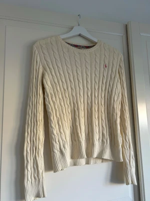 Vit kabelstickad tröja från Ralph Lauren - Kabelstickad tröja i offwhite från Ralph Lauren med rund halsringning och lång ärm. Tröjan har det klassiska rosa Polo-logotypen broderad på bröstet och ribbade muddar vid ärmslut och nederkant🩷🤍