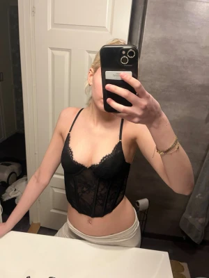 Svart spets korsett/linne med tunna axelband - Säljer en svart korsett/linne i spets med tunna justerbara axelband och v-ringad urringning. Linnet har genomskinliga spetsdetaljer och markerad midja för en snygg siluett. Perfekt för dig som vill ha något stilrent och trendigt. Materialet är troligtvis en blandning av spets och mesh.
