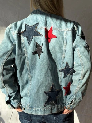 Ljusblå jeansjacka med stjärnor - Jeansjacka med stora dekorativa stjärnpatches i rött och mörkblått på ryggen. Perfekt för dig som vill sticka ut med en unik och trendig look.🩵