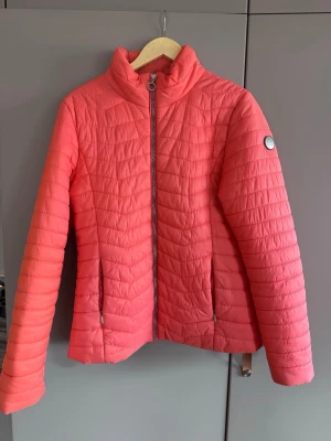 Korallrosa pufferjacka med dragkedja - Snygg korallrosa pufferjacka med hög krage och hel dragkedja framtill. Jackan har två fickor med dragkedja och quiltad design. Märke syns på ärmen. Perfekt för kalla vinterdagar och ger en fräsch look till din outfit.