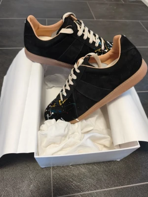 Maison Margiela GATS svart sneakers paint  - Maison Margiela sneakers i svart mocka och läder med unika färgstänk i vitt, gult och rött. Klassisk låg modell med vita snören och beige sula. Snygg och konstnärlig design som sticker ut. Kommer med originalkartong.