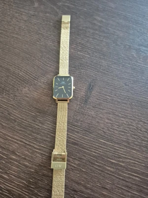 Guldig klocka från Daniel Wellington - Stilren klocka från Daniel Wellington med fyrkantig svart urtavla och guldfärgat mesh-armband. Klockan har minimalistisk design med tunna guldiga visare och index. Perfekt accessoar för dig som gillar klassisk men modern stil. Har tyvärr inte lådan till klockan kvar. 