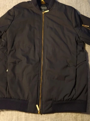 Svart bomberjacka Peak Performance S - Säljer en svart bomberjacka från Peak Performance Anniversary Collection. Jackan har guldfärgade dragkedjor, ribbade muddar och en klassisk ficka på ärmen. Materialet är polyester och fodret är lent och glansigt. Perfekt för dig som vill ha en stilren och tidlös look.