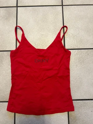 Röd linnetopp från DKNY storlek S - Snygg röd linnetopp från DKNY med tunna axelband och v-ringad hals. Toppen har DKNY-loggan framtill i små glittriga stenar. Perfekt för dig som gillar enkel men ändå statement stil.