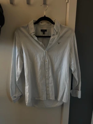 Ljusblå randig skjorta från Gant - Klassisk ljusblå skjorta från Gant med tunna vita ränder och broderad logga på bröstet. Skjortan har lång ärm, knappar framtill och en avslappnad passform. Perfekt till jeans eller chinos för en clean look.