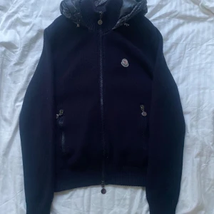 Svart pufferjacka tricot cardigan moncler - Snygg svart pufferjacka från Moncler med stickade ärmar och vadderad rygg samt huva. Jackan har dragkedja framtill, två fickor med knapp och Moncler-logga på bröstet. Perfekt för kalla dagar och riktigt trendig look.