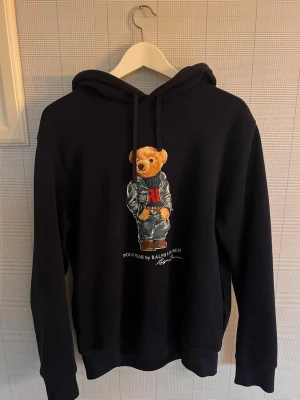 Marin blå  hoodie Polo Ralph Lauren björn - Snygg marinblå hoodie från Polo Ralph Lauren med den ikoniska björnen på bröstet. Hoodien har dragsko i huvan och ribbade muddar vid ärmar och nederkant. Perfekt för dig som gillar streetwear och vill ha något som sticker ut.