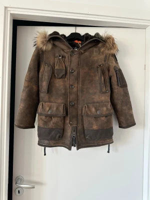 Brun parkas med äkta päls och många fickor - Parajumpers  - Säljer en brun parkas med huva och äkta päls och läderdetalj. Jackan har flera stora och små fickor med dragkedjor och knappar, samt förstärkta partier vid ärmar och nederkant. Snygg vintage-look och perfekt för kalla dagar. Nypris: 10 000 kr Storlek: 128