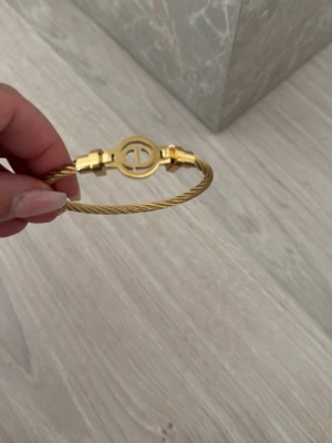 Guldigt kabelarmband med logga - Snyggt armband i guldfärgad metall med tvinnad kabeldesign och en rund logodetalj framtill. Armbandet har en stilren och modern look som passar perfekt till din vardagsstil. Enkel att matcha med andra accessoarer för en trendig vibe.