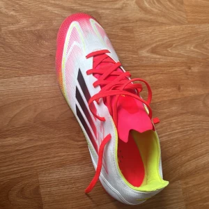 Adidas F50 vita och neon fotbollsskor - Snygga Adidas F50 fotbollsskor i vitt med neonrosa och gula detaljer. Skorna har svarta ränder på sidan, snörning och en lätt ovandel i syntetmaterial. Sulan är rosa med fasta dobbar för bra grepp på planen. Perfekta för dig som vill sticka ut på matchen! Använda 10 gånger och är i använt skick. Priset kan diskuteras vid snabb affär!