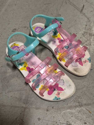 Sandaler - Säljer ett par sandaler från Gios Eppo med transparenta rosa remmar och turkosa spännen. Sulan är vit med färgglada fjärilar tryckta på innersulan. Tillverkade i plastmaterial, perfekta för sommarens alla äventyr. Finns fläckar.