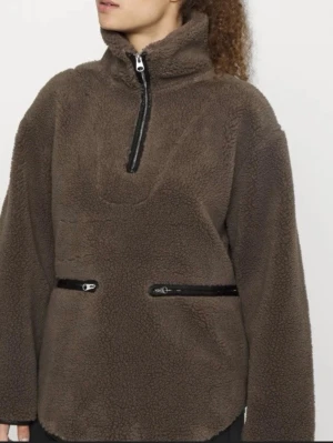 teddy anorak med dragkedjor - Mysig brun teddytröja med hög krage och halv dragkedja framtill. Två svarta dragkedjefickor på magen ger en cool detalj. Perfekt för kalla dagar och riktigt skön att ha på sig. Materialet är fluffigt och värmande.