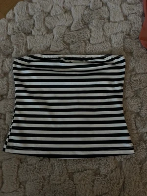 Randig svartvit tubtopp - Säljer en stilren tubtopp med svarta och vita horisontella ränder. Toppen är axelbandslös och har en tight passform som sitter snyggt på kroppen. Perfekt att matcha med jeans eller kjol för en trendig look.