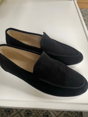 Svarta mockaloafers med vit sula - Snygga svarta loafers i mocka med vit sula och beige innersida. Skorna har en klassisk, stilren design utan snörning. Materialet ger en mjuk känsla och den vita sulan ger en modern touch. Endast testade men försmå! Hör av dig vid eventuella frågor