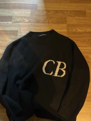Svart stickad tröja Cole Buxton - Svart stickad tröja från Cole Buxton med stora vita bokstäver 'CB' på bröstet. Tröjan har rund halsringning och ribbade muddar vid ärmslut och nederkant. Perfekt för en avslappnad och trendig stil.