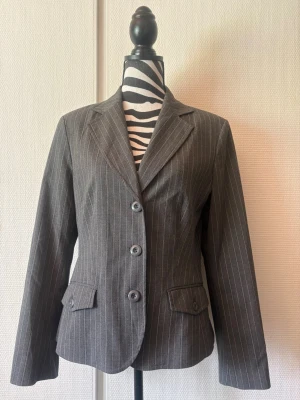 Grå kavaj med smala ränder, size M - Snygg grå kavaj med smala vita kritstrecksränder och tre knappar framtill. Klassisk krage, figursydd passform och två fickor med lock. Perfekt för en stilren och trendig look. Materialet känns som en blandning av bomull och polyester.