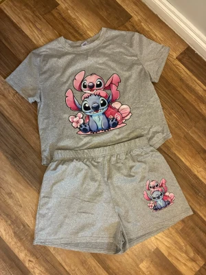 Grå pyjamas med Stitch & Angel-motiv - Supermysig grå pyjamas med kortärmad t-shirt och shorts. Båda delarna har ett färgglatt tryck av Stitch och Angel från Disney. Materialet är mjukt och stretchigt, perfekt för chillkvällar hemma. Elastisk midja på shortsen för extra komfort.