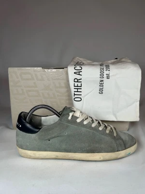 Gråa sneakers från Golden Goose - Storlek 39 men passar 40 Snygga gråa sneakers från Golden Goose med ovandel i mocka och vita skosnören. Skorna har en låg silhuett och svart detalj på hälen med vit text. Yttersulan är vit med en något sliten look. Perfekta för en avslappnad och trendig stil.