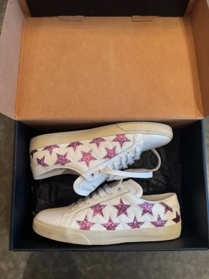 Saint Laurent sneakers med rosa stjärnor - Vita sneakers från Saint Laurent med glittriga rosa stjärnor längs sidorna. Skorna har klassisk låg profil, vita skosnören och beige sula. Tillverkade i läder med coola detaljer som sticker ut och ger en unik vibe.