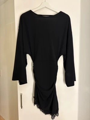 Svart långärmad klänning med spets - Svart långärmad klänning med draperad halsringning och asymmetrisk nederkant dekorerad med spets. Klänningen har en elegant och stilren look, perfekt för dig som gillar en klassisk men ändå unik stil. Materialet känns mjukt och följsamt.