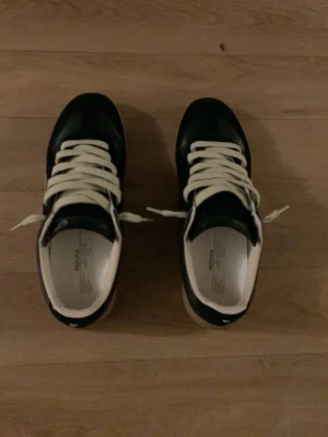 Svarta Maison margiela gats - Snygga svarta sneakers från maison margiela  med vita detaljer och vita snören. Skorna har en klassisk låg profil och är tillverkade i skinn med en vit innersula och beige yttersula. Perfekta för dig som gillar stilrena och tidlösa sneakers.
