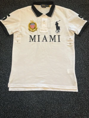 Vit Polo Ralph Lauren MIAMI t-shirt - STORLEK M (SLIM FIT) Vit skjorta med svart krage och ärmslut. Framsidan har broderad logga, emblem och texten MIAMI. Baksidan har en stor svart siffra 5. Klassisk pikéstil med knappar vid halsen och detaljer på sidorna. BARA ANVÄNTS 1 GÅNG❗️