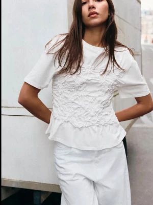 Vit blus med volang och struktur - Säljer en vit blus med korta ärmar och volang nedtill. Toppen har ett strukturerat, rynkat parti framtill som ger en unik look. Perfekt att styla med jeans eller kjol för en trendig outfit. Ny med prislapp.