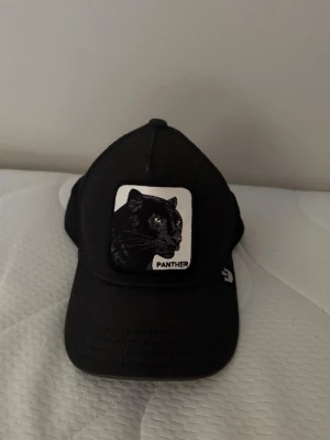 Svart truckerkeps med pantermärke - Säljer en svart truckerkeps från Goorin Bros med ett broderat patch framtill som visar en panter och texten 'Panther'. Kepsen har mesh baktill för extra ventilation och justerbar snapback. Perfekt för dig som gillar djurmotiv och streetwear.