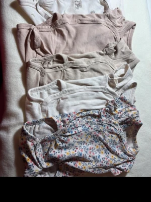 6-pack neutrala och blommiga bodys - Säljer ett 6-pack bodys i mjuk bomull. Färgerna är beige, vitt och ljusrosa, samt en body med blommigt mönster i pastellfärger. Några har diskreta ränder och en har små djurmotiv. Alla har tryckknappar vid axel och gren för enkel påklädning.