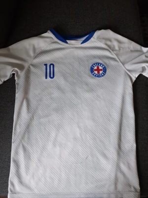 Vit England fotbollströja nr 10 - Säljer en vit fotbollströja med blå detaljer och England-märke på bröstet. Tröjan har nummer 10 tryckt både fram och bak samt texten 'ENGLAND' på ryggen. Tillverkad i ett lätt och ventilerande material, perfekt för träning eller match.