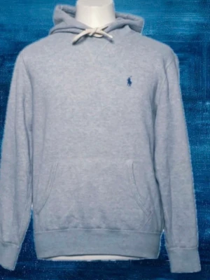 Grå hoodie från Polo Ralph Lauren - Snygg ljusgrå hoodie från Polo Ralph Lauren med klassisk blå logga broderad på bröstet. Hoodien har huva med vita dragsnören och en stor känguruficka framtill. Tillverkad i mjukt bomullsmaterial och har ribbade muddar vid ärmslut och nederkant.
