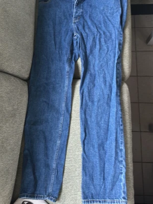 Blå raka jeans i klassisk modell - Säljer ett par blå jeans med rak passform och hög midja. Jeansen har fem fickor och stängs med dragkedja och knapp. Tillverkade i klassisk denim, perfekta för en avslappnad stil. Inga synliga loggor eller märken.