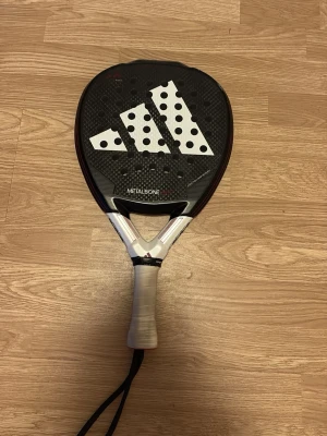 Adidas Metalbone padelracket - Säljer ett Adidas Metalbone padelracket i svart med stora vita detaljer och prickar. Racketen har diamantformad design, grepp i vitt och handledsrem. Tillverkat i kolfiber för extra hållbarhet och prestanda. Perfekt för dig som vill ta ditt padelspel till nästa nivå.