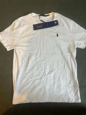 Vit t-shirt från Polo Ralph Lauren - Klassisk vit t-shirt från Polo Ralph Lauren med den ikoniska lilla loggan broderad på bröstet. T-shirten har rund hals, korta ärmar och är gjord i mjuk bomull för en skön känsla. Perfekt basplagg till garderoben.