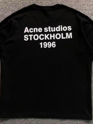 Svart Acne Studios t-shirt - Svart t-shirt från Acne Studios med vit text 'Acne Studios STOCKHOLM 1996' på bröstet. Klassisk rund hals och korta ärmar. Perfekt för en stilren och trendig look.
