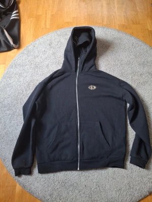 Svart zip hoodie från Black Squad - Svart hoodie med dragkedja från Black Squad i relaxed fit. Klassisk huva, två fickor framtill och diskret broderad logga på bröstet. Mjuk insida som känns skön mot huden. Perfekt för en chill och avslappnad stil.