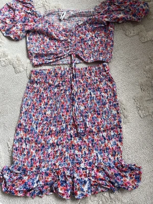 Blommigt set med puffärm och volang - Säljer ett färgglatt tvådelat set med blommönster i rosa, blått, rött och vitt. Toppen har puffärmar, knytning framtill och smockad resår. Kjolen är kort med smockad midja och volangkant. Perfekt för dig som gillar lekfulla och trendiga plagg!