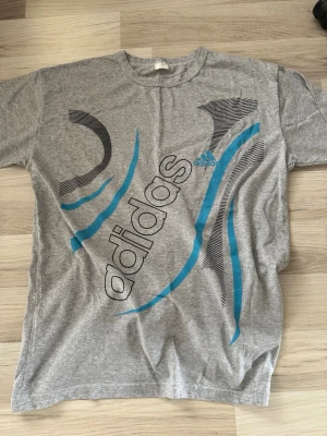 Grå Adidas t-shirt med blå detaljer - Säljer en grå t-shirt från Adidas med stora grafiska tryck i svart och blått på framsidan. T-shirten har rund halsringning och korta ärmar. Perfekt för en sportig och avslappnad stil. Materialet känns mjukt och skönt, troligtvis bomull.