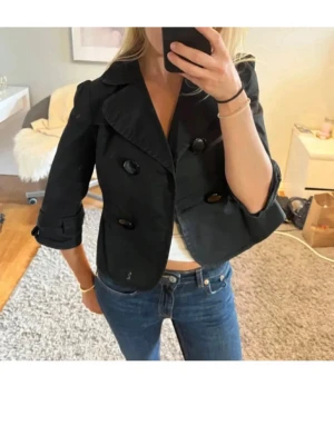 Vintage trenchcoat  - Tänkte kolla intresset på denna. Snygg vintage trenchcoat med dubbelknäppning och stora knappar. Jackan har trekvartsärmar med dekorativa detaljer vid ärmslut och bred krage. Perfekt för dig som vill ha en trendig och stilren look.
