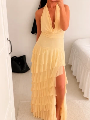 Ljusgul långklänning med volanger - Säljer en ljusgul långklänning med djup v-ringning och halterneck. Klänningen har flera lager volanger och en hög slits framtill som ger en snygg och trendig look. Perfekt för sommarens alla tillfällen!