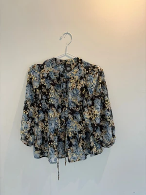 Blommig blus med knytband  - Snygg svart blus med blåa och guldiga blommönster. Blusen har långa ärmar med resår vid ärmslut och knytband i midjan. Tillverkad i ett tunt, lätt material som ger ett luftigt intryck. Perfekt för dig som gillar fina blommiga mönster. 