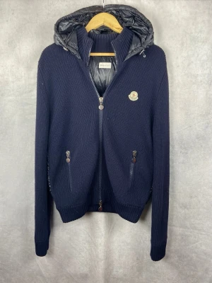 Mörkblå stickad jacka från Moncler - Mörkblå stickad jacka från Moncler med avtagbar huva i blankt material och två dragkedjeförsedda fickor. Jackan har ribbade muddar, Moncler-logga på bröstet och hel dragkedja framtill. Perfekt för kyliga dagar.