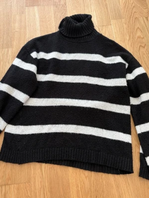 Svartvit randig polotröja stickad - Stickad svart tröja med breda vita horisontella ränder och hög polokrage. Tröjan har en avslappnad passform, långa ärmar och ribbade muddar vid ärmslut och nederkant. Perfekt för dig som gillar klassiska mönster och vill ha något mysigt.
