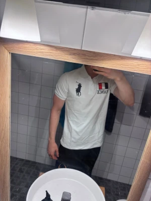 Vit pikétröja Polo Ralph Lauren - Vit pikétröja från Polo Ralph Lauren med klassisk krage och knappar. Broderad logga i svart på bröstet, FRANCE-text och fransk flagga på vänster sida samt en stor svart siffra 3 på ärmen.