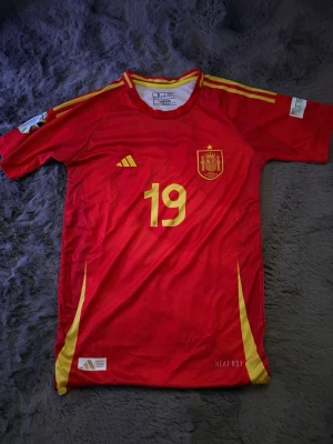 Spanien hemmatröja 2024 lamine yamal - Röd Spanien matchtröja från Adidas med gula detaljer och nummer 19. På bröstet finns landslagets emblem och Adidas-logga, samt texten 'HEAT.RDY' för materialet. På ryggen står Lamine Yamal och nummer 19 i gult. Korta ärmar och klassisk sportig passform.