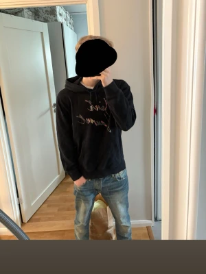 Svart Saint Laurent hoodie - Säljer en svart hoodie från Saint Laurent med färgglad logga framtill. Hoodien har huva med dragsko och en stor magficka. Tillverkad i mjukt bomullsmaterial och har ribbade muddar vid ärmslut och nederkant. Perfekt för en avslappnad och trendig stil. Köpt på plick för 1500kr. 