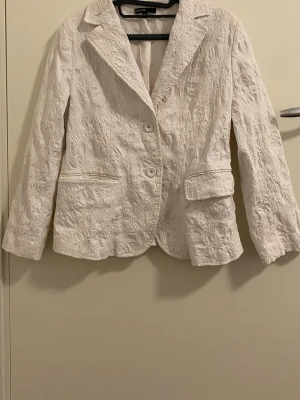 Vit kavaj med struktur från KappAhl - Snygg vit kavaj från KappAhl med unik strukturerad yta. Klassisk krage, två knappar framtill och fickor med lock. Perfekt för dig som vill sticka ut med en stilren men annorlunda look. Materialet känns lätt och har en cool textur.
